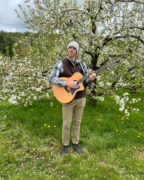 Theo Manazir standing by a tree holding a guitar and smiling